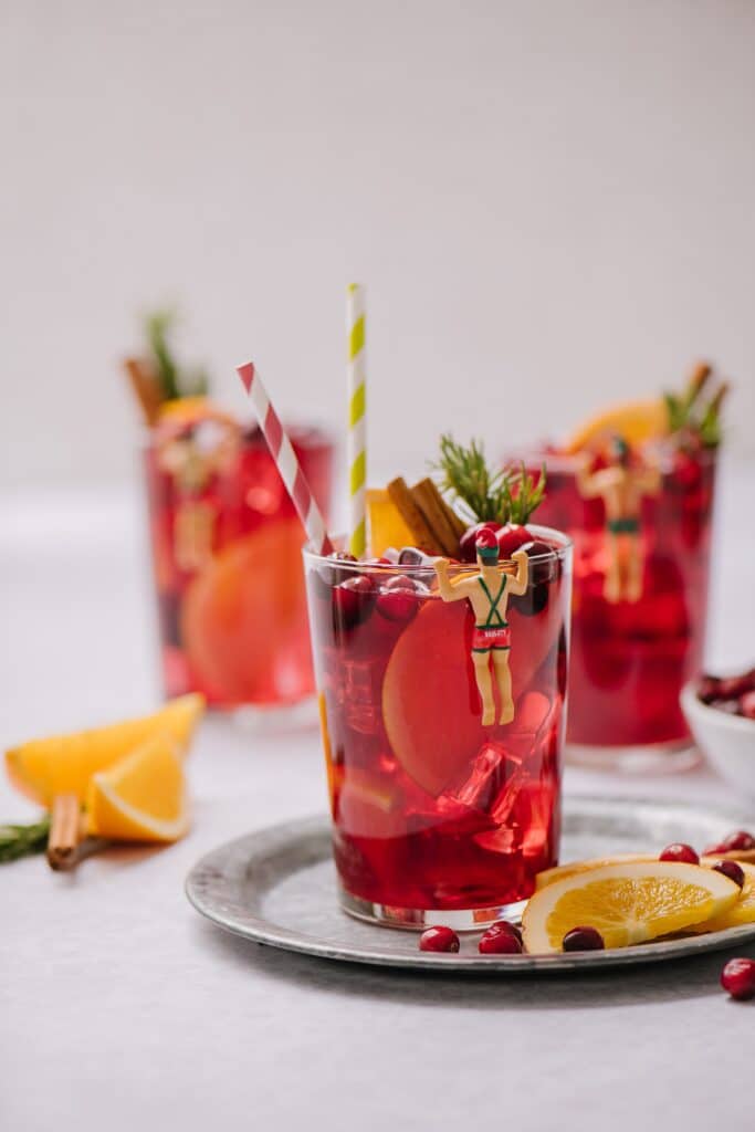 A glass of mulled red wine sangria on ice, garnished with cinnamon sticks, rosemary, and a straw. There is also a drink marker that looks like a swimmer with a Santa hat on.