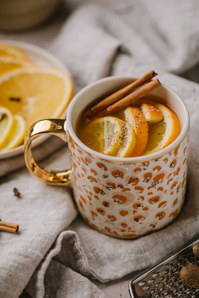 A traditional warm spiced apple cider, topped with cinnamon sticks and oranges. Sliced oranges on a plate are in the background.